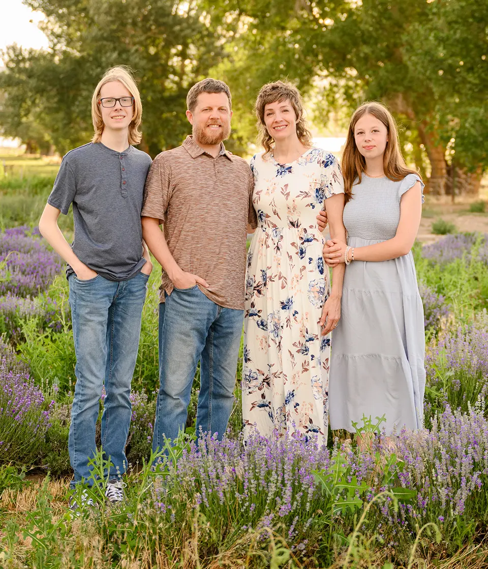 Family of four standing together outdoors near purple flowers and trees.