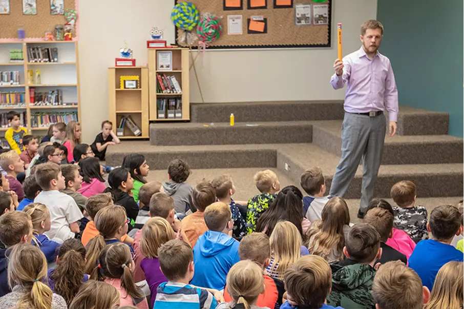 Man presenting to a classroom full of children seated on the floor.