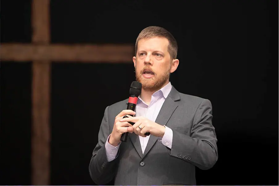 Speaker in a suit holding a microphone on a dark stage.