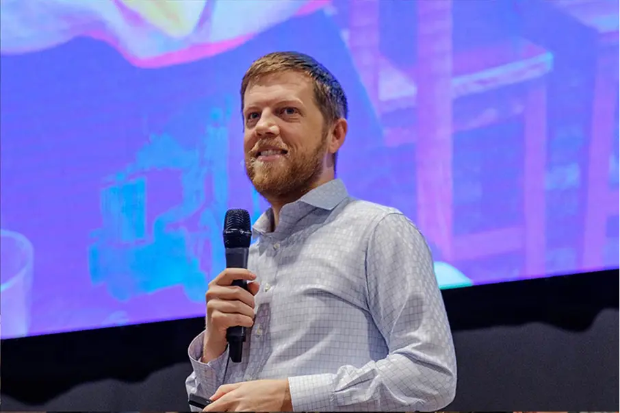 Man speaking on stage with a purple-lit screen behind him.