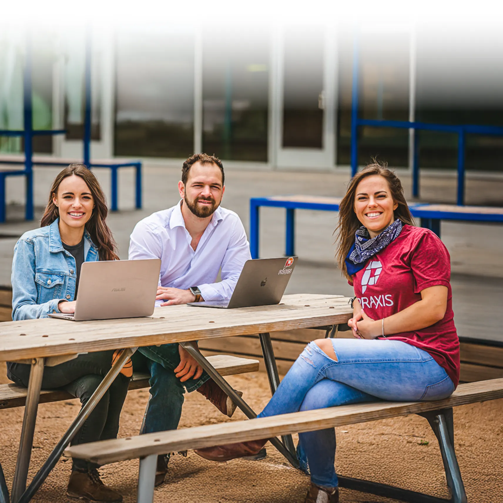 Three young adults sitting at a picnic table with laptops outside.